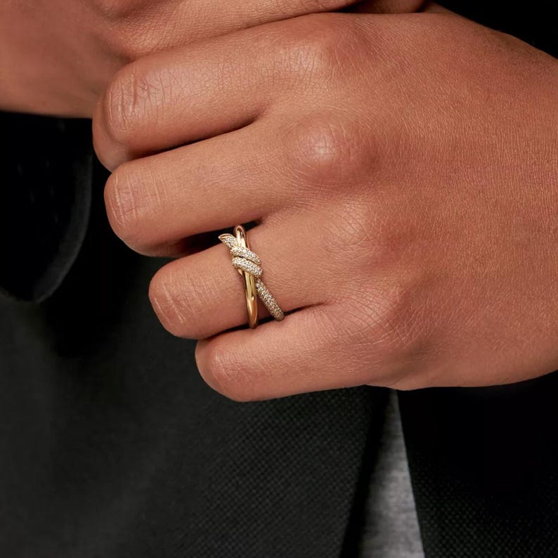 K&ouml;nnte beinhalten: Ein goldfarbener Ring mit einem Knoten-Design, besetzt mit kleinen, funkelnden Steinen. Der Ring wird an einem Finger getragen und zeigt seine komplizierten Details und seinen eleganten Stil. Das Design des Rings ist eine moderne Interpretation eines klassischen Knotens.