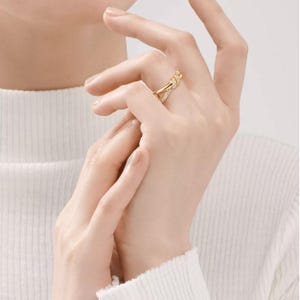 K&ouml;nnte beinhalten: Ein goldfarbener Ring mit einem Knoten-Design, verziert mit kleinen, funkelnden Steinen. Der Ring wird an einer Hand getragen, wobei die Finger sanft gekr&uuml;mmt sind. Der Hintergrund ist in einem weichen, neutralen Farbton gehalten.