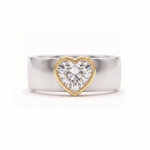 May include: A wide silver band ring featuring a central heart-shaped clear gemstone set within a gold-toned bezel. The band has a smooth, polished finish, and the heart-shaped stone is faceted, reflecting light from its center point outward.