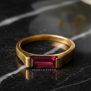 Puede incluir: Un anillo de oro con una gema rectangular de color rojo intenso. El anillo tiene un diseño simple y moderno con un acabado liso y pulido. El texto "FLORA & FORGE" es visible en el anillo.