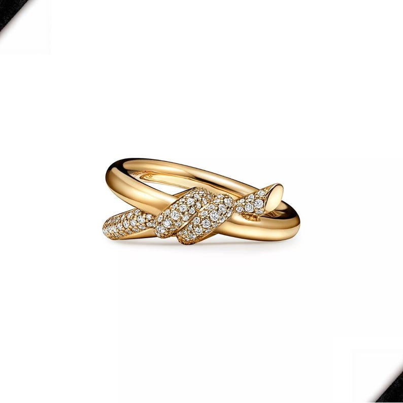 K&ouml;nnte beinhalten: Ein goldfarbener Ring mit Knoten-Design. Der Knoten ist mit kleinen, klaren Edelsteinen besetzt. Der Ring hat eine polierte, reflektierende Oberfl&auml;che und ist vor wei&szlig;em Hintergrund platziert. Ein Schmuckst&uuml;ck.