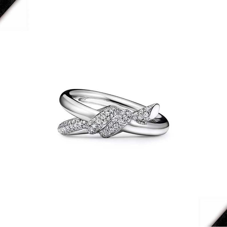 K&ouml;nnte beinhalten: Ein silberfarbener Ring mit Knoten-Design. Der Knoten ist mit kleinen, funkelnden Edelsteinen besetzt. Der Ring hat eine polierte, reflektierende Oberfl&auml;che, vor wei&szlig;em Hintergrund.