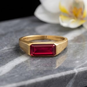 Puede incluir: Un anillo dorado con una gema rectangular de color rojo intenso. El anillo tiene un acabado de metal cepillado y un diseño clásico y minimalista. La gema está engastada en un bisel. El anillo se muestra sobre una superficie gris.