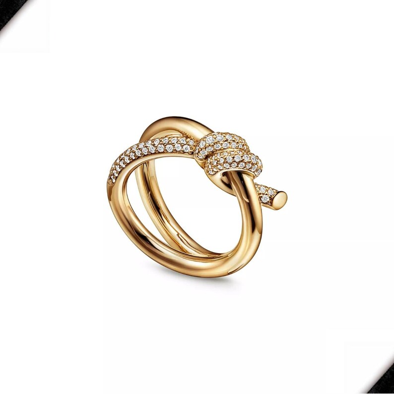 K&ouml;nnte beinhalten: Ein goldfarbener Ring mit einem Knotendesign. Der Knoten ist mit kleinen, funkelnden Steinen besetzt. Der Ring hat eine glatte, polierte Oberfl&auml;che und einen klassischen, eleganten Stil. Der Ring hat die Gr&ouml;&szlig;e 54 (EU).