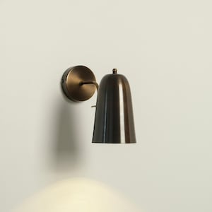May include: A bronze-colored wall sconce with a conical shade and a circular mounting plate. The sconce is designed to be mounted on a wall and emits a downward light. The fixture has a classic, minimalist design.