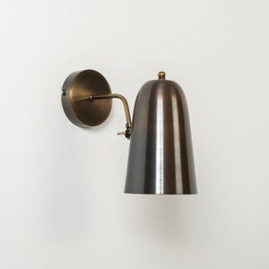 May include: A bronze-finished wall sconce. It has a circular base, a curved arm, and a bell-shaped shade. The design is reminiscent of vintage or industrial lighting fixtures, suitable for interior use.