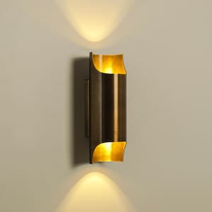 Op de afbeelding: Een bronzen, cilindrische wandlamp met een gebogen boven- en onderkant. De lamp straalt een warm, goudkleurig licht naar boven en beneden uit, waardoor een zachte gloed ontstaat. De armatuur is aan een neutraal gekleurde muur bevestigd.