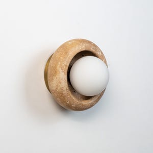 May include: A modern wall sconce featuring a beige, circular stone design with a white spherical light. The sconce has a brass-colored base and is mounted on a white surface. The light fixture is a contemporary design.