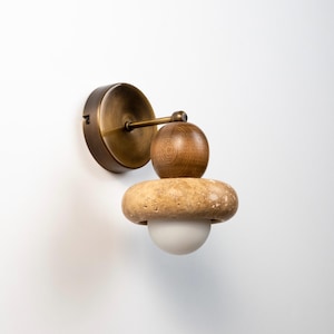 May include: A modern wall sconce with a brass finish, featuring a wooden sphere, a stone-like ring, and a white globe light. The fixture is mounted on a white wall, showcasing a minimalist design.