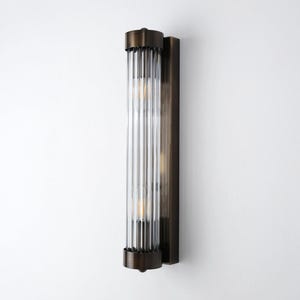 May include: A vertical wall sconce with a bronze finish. The sconce features a clear, ribbed glass tube that houses two light bulbs. The fixture is mounted on a rectangular bronze backplate, creating a modern and elegant lighting solution.