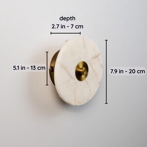 May include: A circular wall sconce featuring a marble face and brass detailing. The sconce measures 20 cm high, 13 cm wide, and 7 cm deep. The marble has a cream and tan pattern, and the brass has a warm, metallic finish.