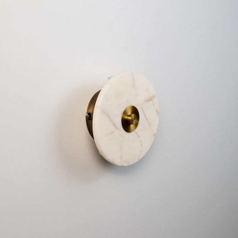 May include: A circular wall sconce made of white marble with gold veining and a brass centre. It is mounted on a brass base. This decorative lighting fixture is designed for wall mounting. The diameter is approximately 15 cm.