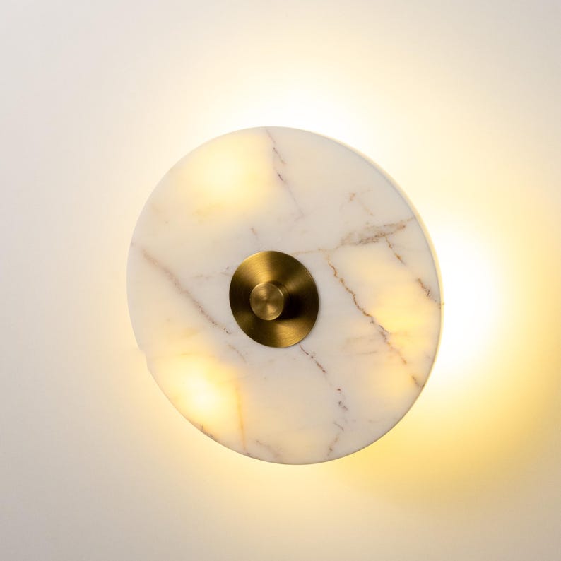 May include: A circular wall light made of white marble with brown veining and a brass centre. The light is illuminated, emitting a warm glow. The design is modern and minimalist, suitable for interior lighting.