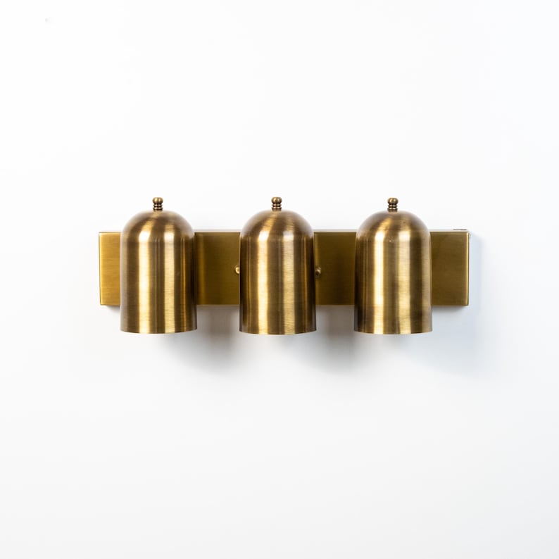 May include: A wall-mounted light fixture featuring a rectangular brass backplate and three cylindrical, dome-topped brass shades. Each shade has a smooth, brushed metallic finish and a small spherical finial at the very top of the dome.