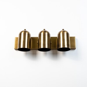 May include: A three-light wall fixture featuring a rectangular brass backplate and three cylindrical, dome-topped brass shades. Each shade is oriented vertically, creating a uniform row of metallic lighting components with a brushed finish.