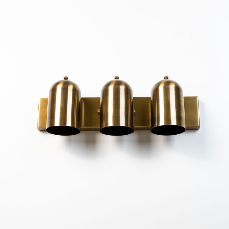 May include: A three-light wall fixture featuring a rectangular brass backplate and three cylindrical, dome-topped brass shades. Each shade is oriented vertically, creating a uniform row of metallic lighting components with a brushed finish.