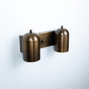 May include: A wall-mounted light fixture featuring a rectangular backplate in a brushed bronze finish. Two cylindrical, dome-topped shades in the same metallic bronze finish are attached to the plate, each topped with a small decorative knob.