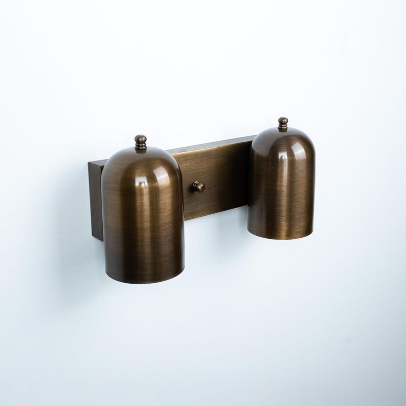 May include: A wall-mounted light fixture featuring a rectangular backplate in a brushed bronze finish. Two cylindrical, dome-topped shades in the same metallic bronze finish are attached to the plate, each topped with a small decorative knob.