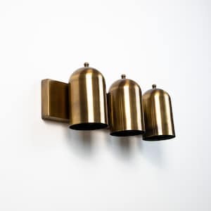 May include: A wall-mounted light fixture featuring three dome-shaped shades in a brushed brass finish. Each shade is attached to a rectangular backplate and topped with a small decorative finial, creating a uniform and linear lighting arrangement.