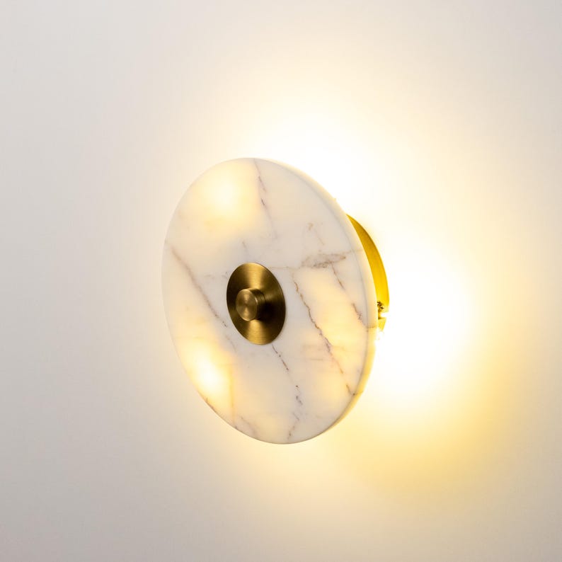 May include: A circular wall light with a white marble front and brass detailing. The marble features delicate grey veining. The light is on, emitting a warm glow. The design is modern and minimalist, suitable for various interiors.
