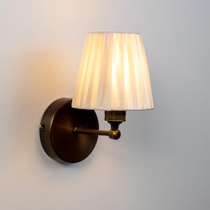 Op de afbeelding: Een wandlamp met een ronde, donkerbronzen voet en een bijpassende horizontale arm. De lamp is voorzien van een kleine, gebroken witte, geplooide stoffen kap die een warm licht uitstraalt en is bevestigd aan een metalen fitting.