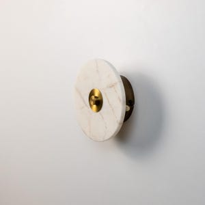 May include: A circular wall sconce made of white marble with gold and brass details. The sconce has a gold central element and a brass backing. The design is minimalist and modern, suitable for interior decor.