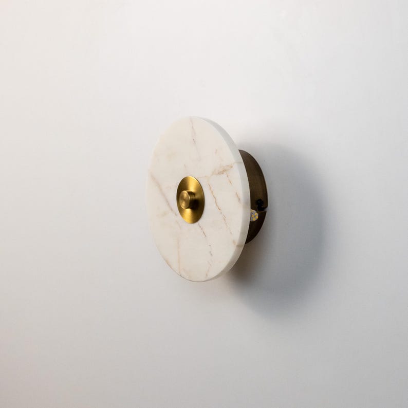May include: A circular wall sconce made of white marble with gold and brass details. The sconce has a gold central element and a brass backing. The design is minimalist and modern, suitable for interior decor.
