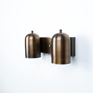 May include: A wall-mounted light fixture featuring two dome-shaped shades with a brushed bronze finish. The shades are attached to a rectangular backplate, each topped with a small decorative finial. The metal surface has a warm, metallic sheen.