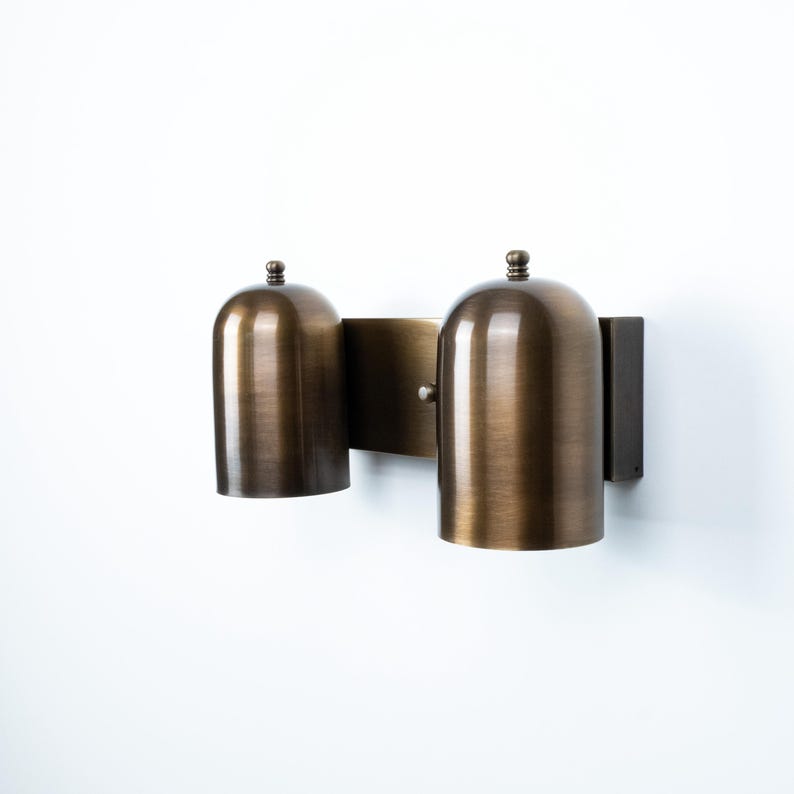 May include: A wall-mounted light fixture featuring two dome-shaped shades with a brushed bronze finish. The shades are attached to a rectangular backplate, each topped with a small decorative finial. The metal surface has a warm, metallic sheen.