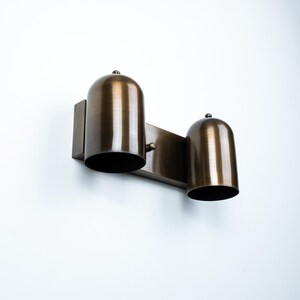 May include: A wall-mounted light fixture featuring two dome-shaped shades with a brushed bronze finish. The fixture consists of a rectangular backplate connected to a horizontal bar that supports the two downward-facing cylindrical metal shades.