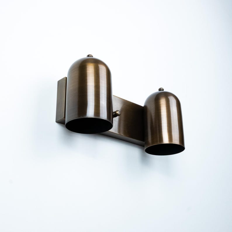 May include: A wall-mounted light fixture featuring two dome-shaped shades with a brushed bronze finish. The fixture consists of a rectangular backplate connected to a horizontal bar that supports the two downward-facing cylindrical metal shades.