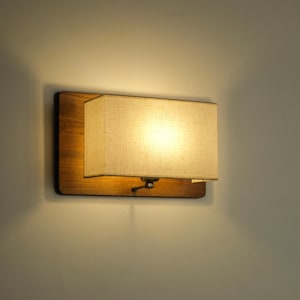 May include: A rectangular wall sconce with a wooden base and a beige fabric shade. The light is on, illuminating the shade. The sconce is mounted on a light-colored wall, creating a warm ambiance.