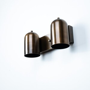 May include: A wall-mounted light fixture featuring two dome-shaped shades with a brushed bronze finish. The fixture consists of a rectangular backplate that supports two cylindrical, downward-facing metal housings, each topped with a small knob.
