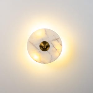 May include: A circular wall light made of white marble with a brass centre. The light is illuminated from behind, producing a warm, ambient glow. The marble has delicate grey veining.