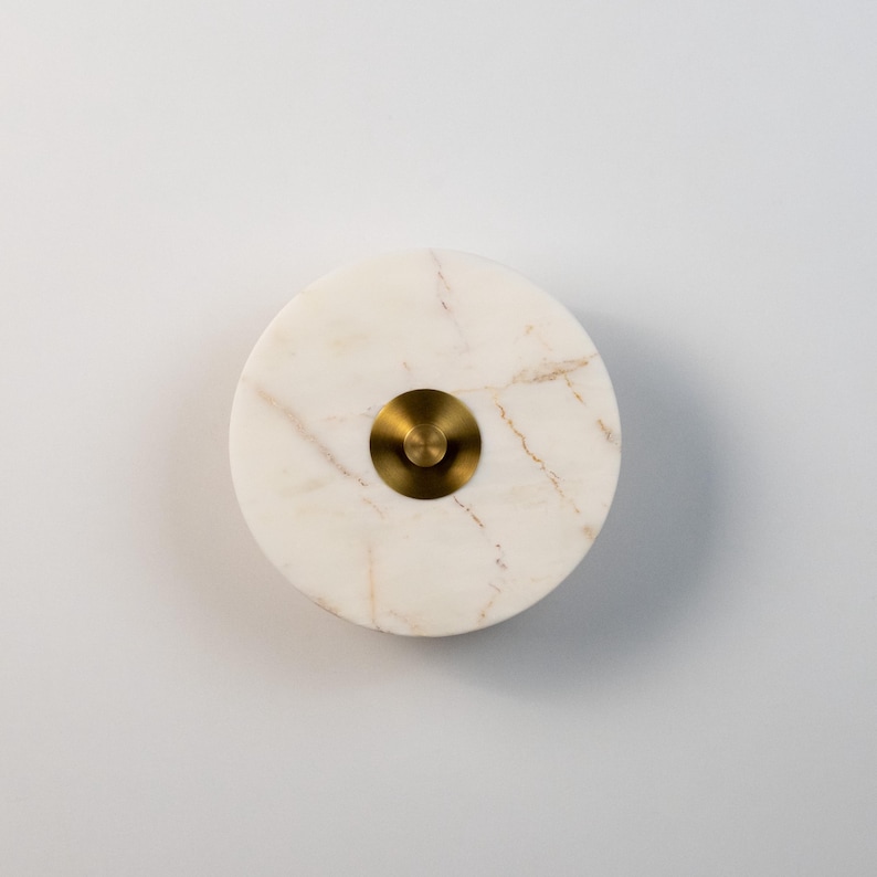 May include: A circular object made of white marble with gold veining, featuring a central brass disc. The marble has a polished surface, and the brass has a brushed finish. The design is minimalist and elegant.