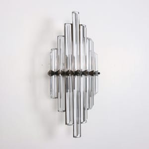Op de afbeelding: Een heldere glazen wandlamp met een geometrisch ontwerp. De lamp heeft meerdere rechthoekige glazen staven die in een trapsgewijs, verticaal patroon zijn gerangschikt. Een donkere metalen band met decoratieve elementen omcirkelt het midden. Gemonteerd op een witte muur.
