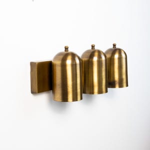 May include: A wall-mounted light fixture featuring three cylindrical brass shades with rounded tops. Each shade is attached to a rectangular brass backplate. The metal has a brushed finish, creating a warm, metallic appearance across the unit.