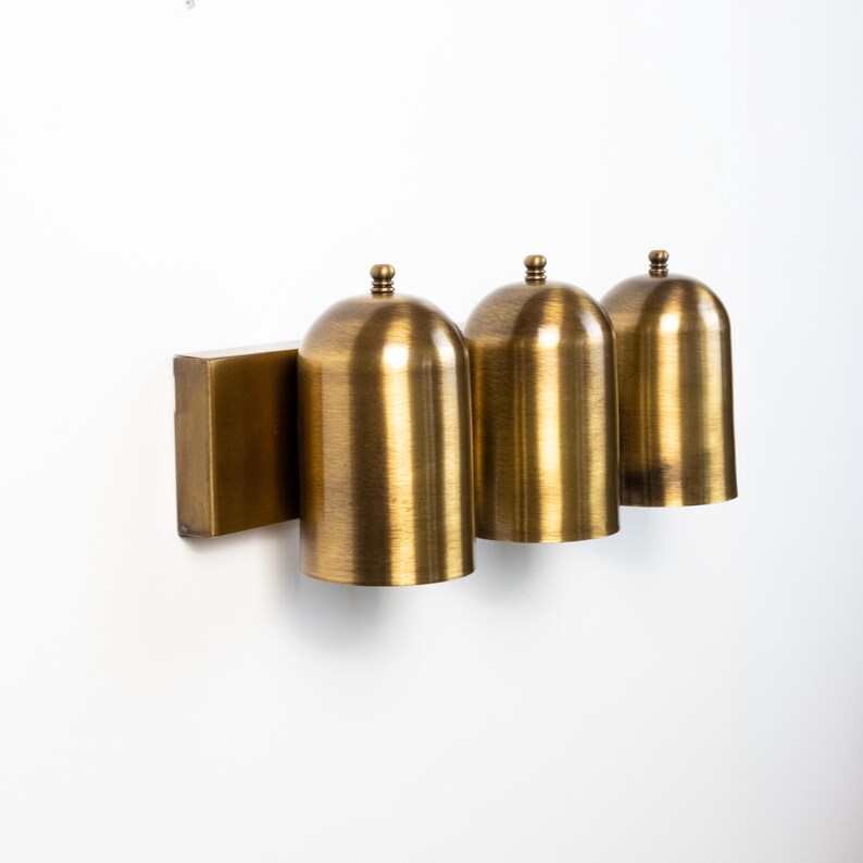 May include: A wall-mounted light fixture featuring three cylindrical brass shades with rounded tops. Each shade is attached to a rectangular brass backplate. The metal has a brushed finish, creating a warm, metallic appearance across the unit.