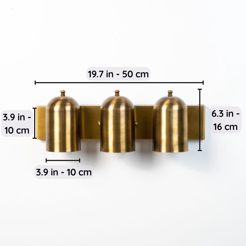 May include: A three-light wall fixture in a brushed brass finish with dome-shaped shades. The fixture measures 19.7 inches wide by 6.3 inches high. Each shade is 3.9 inches wide and 3.9 inches high, mounted on a rectangular backplate.