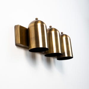 May include: A wall-mounted light fixture featuring three dome-shaped shades made of brushed brass metal. Each shade has a smooth, rounded top with a small decorative finial and a wide, open bottom, all attached to a rectangular brass backplate.