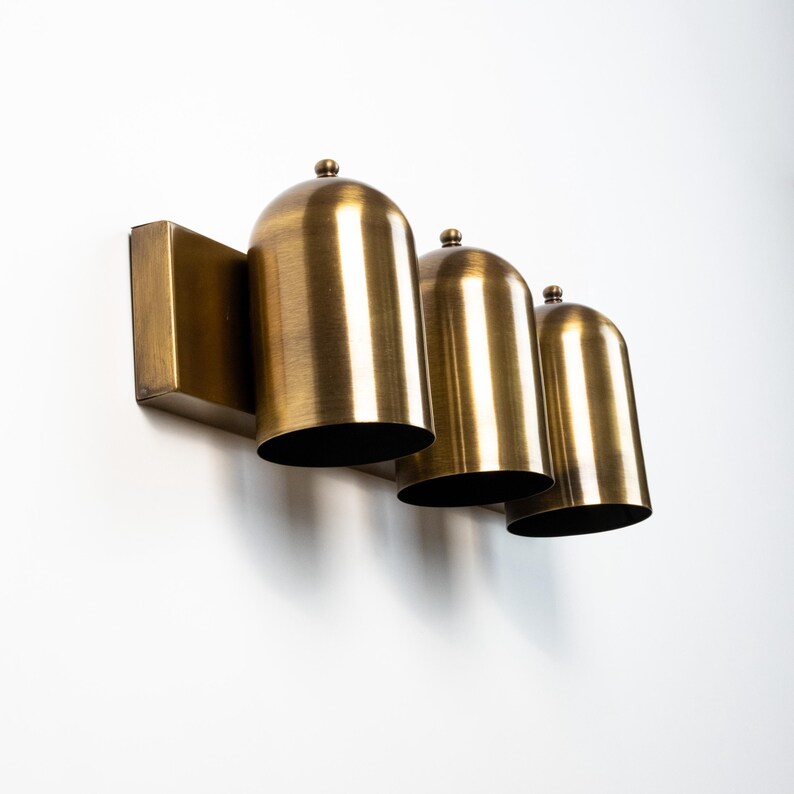May include: A wall-mounted light fixture featuring three dome-shaped shades made of brushed brass metal. Each shade has a smooth, rounded top with a small decorative finial and a wide, open bottom, all attached to a rectangular brass backplate.