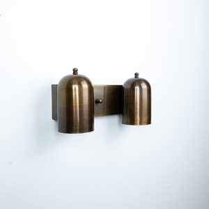 May include: A wall-mounted light fixture featuring a rectangular backplate in a brushed bronze finish. Two dome-shaped shades extend from the plate, each topped with a small decorative finial. The metal surface displays a subtle, brushed texture.