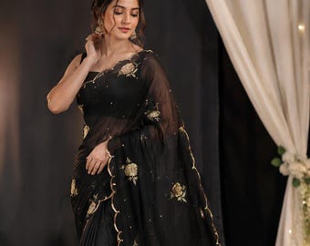 May include: A black saree featuring sheer fabric with gold floral embroidery and a scalloped gold border. The garment includes scattered gold sequins throughout the material, creating a detailed pattern across the length of the draped fabric.