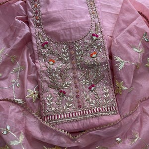 May include: A light pink salwar kameez set. The top has detailed embroidery with silver and gold thread, sequins, and small pink and orange floral details. The dupatta and top feature gold floral embroidery.