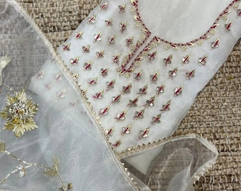 Puede incluir: Una prenda de organza blanca transparente con intrincados bordados en hilos dorados y rojo oscuro. La tela presenta motivos repetitivos en forma de hoja y un adorno floral prominente, rematado con un borde decorativo en el contorno.