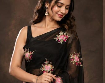 May include: A black saree featuring a sleeveless blouse and a sheer fabric drape adorned with embroidered floral motifs in shades of pink, green, and gold. The embroidery is distributed across the fabric, highlighting the dark base color.