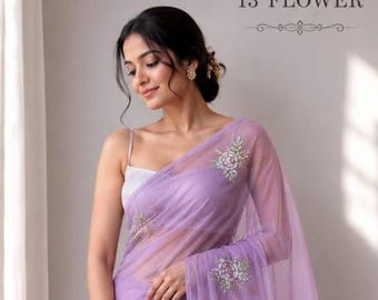 May include: A sheer lavender saree featuring delicate white floral embroidery scattered across the fabric. The garment is paired with a simple white sleeveless blouse. The text 13 FLOWER is visible in the upper right corner of the image.