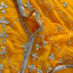 May include: Close-up of a vibrant yellow fabric with intricate silver embellishments. The design features silver floral and geometric shapes, and a silver trim along the edges. The fabric is draped, creating soft folds and shadows.