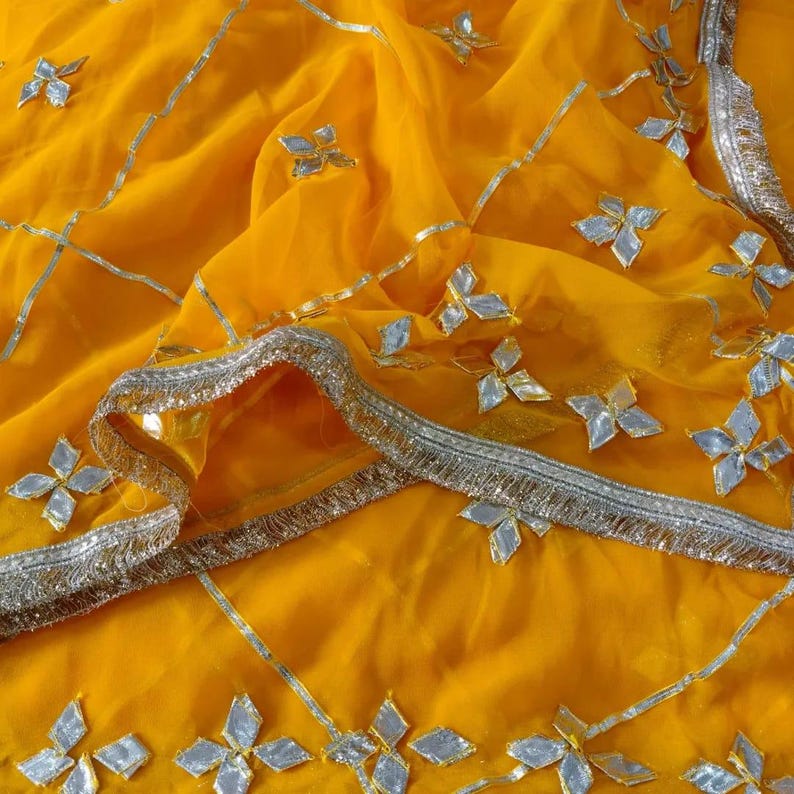 May include: Close-up of a vibrant yellow fabric with silver embellishments. The fabric features a delicate, sheer texture with silver floral and geometric patterns. A silver trim borders the edge, adding a touch of elegance.