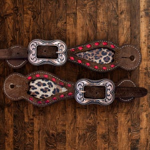 May include: Two brown leather spur straps featuring an ornate silver-toned metal buckle, a central cutout inlay with a leopard print pattern, and decorative red stitching along the edges. The straps have rounded ends with circular holes.